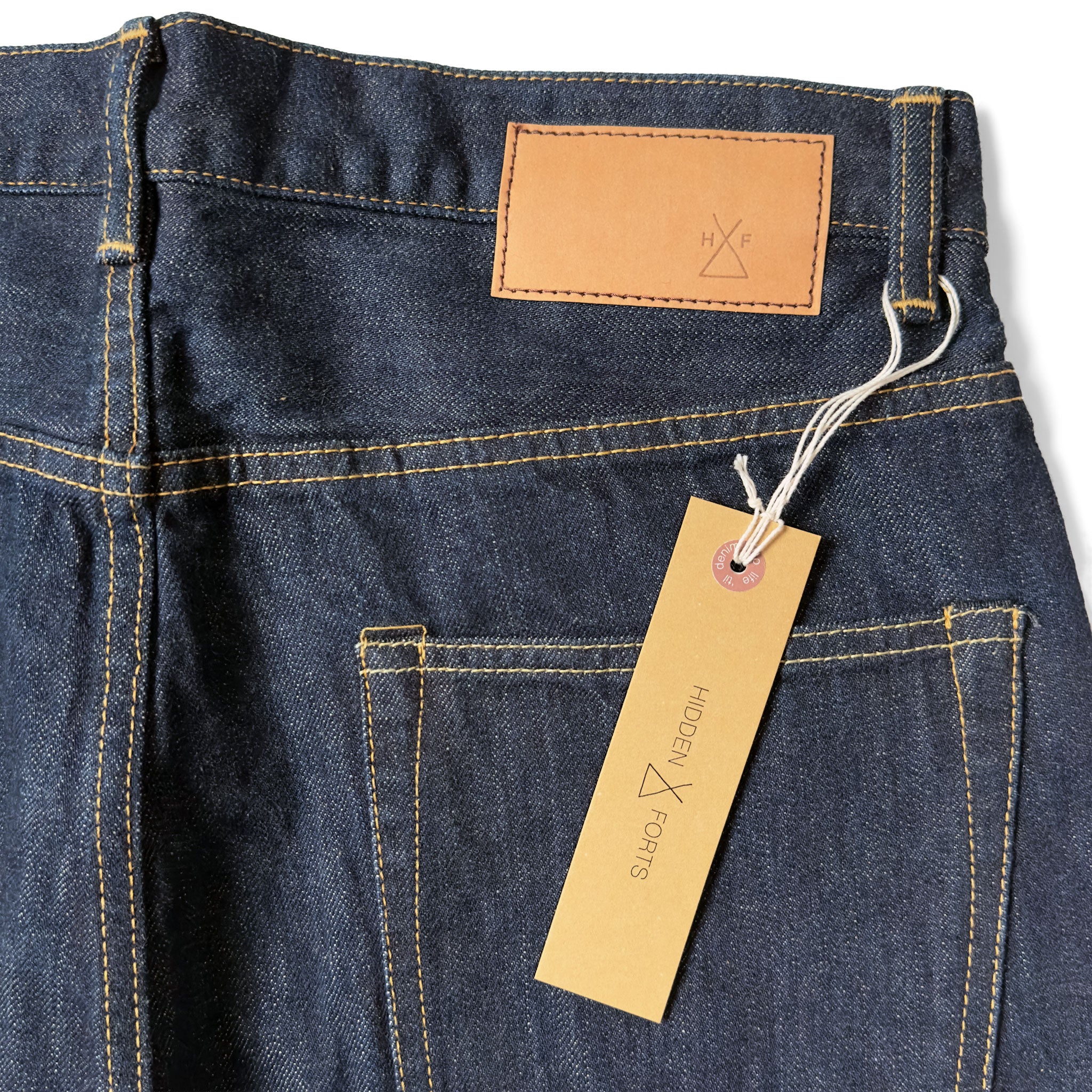 Closeup of the HFD-02 jacron paper patch and kraft paper hangtag with logo on Japanese selvedge  once washed denim relaxed fit tight tapered leg jeans.